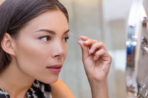 Eine asiatische Frau, die ihre Augenbrauen mit einer Pinzette zupft, verwendet eine Augenbrauenpinzette zu Hause in einem Schminkspiegel im Badezimmer.  Machen Sie sich attraktiver, Nahaufnahme des Gesichts eines Mädchens, während Sie ihre Gesichtsbehaarung entfernen
