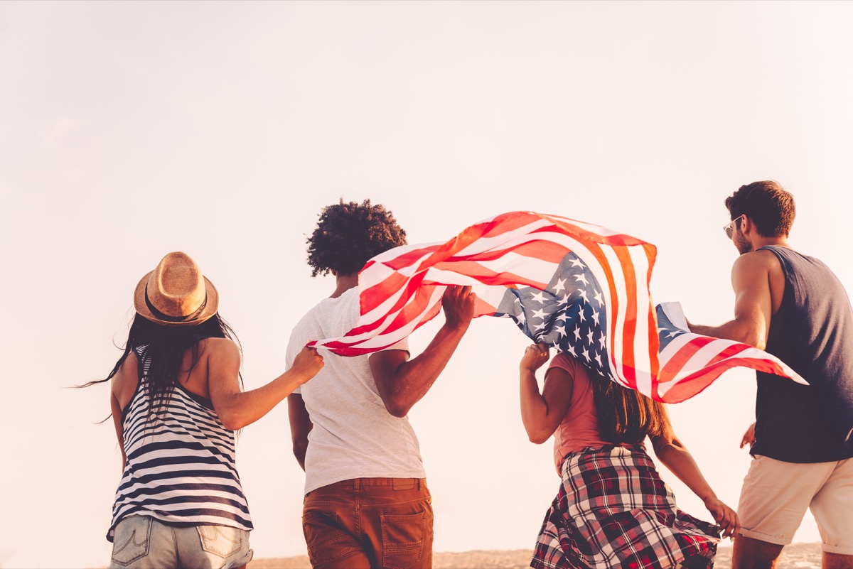 Friends with American flag. Rear view of four young people carrying american flag while running outdoors - Image