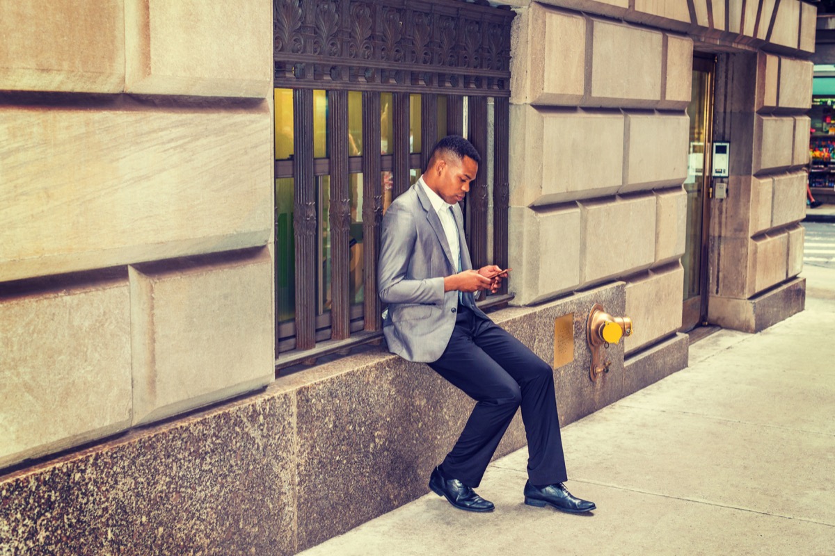 African American man reading, texting on cell phone on street, traveling, working in New York, wearing gray blazer, black pants, leather shoes, sitting on vintage window frame. Instagram filtered look - Image