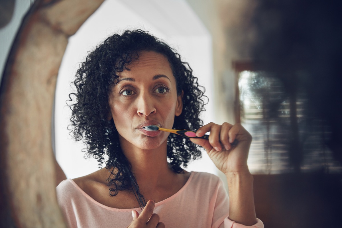 Woman looking in the mirror and brushing her teeth, ways you're damaging teeth