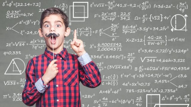 Little boy with fake mustache standing in front of chalkboard with math equations