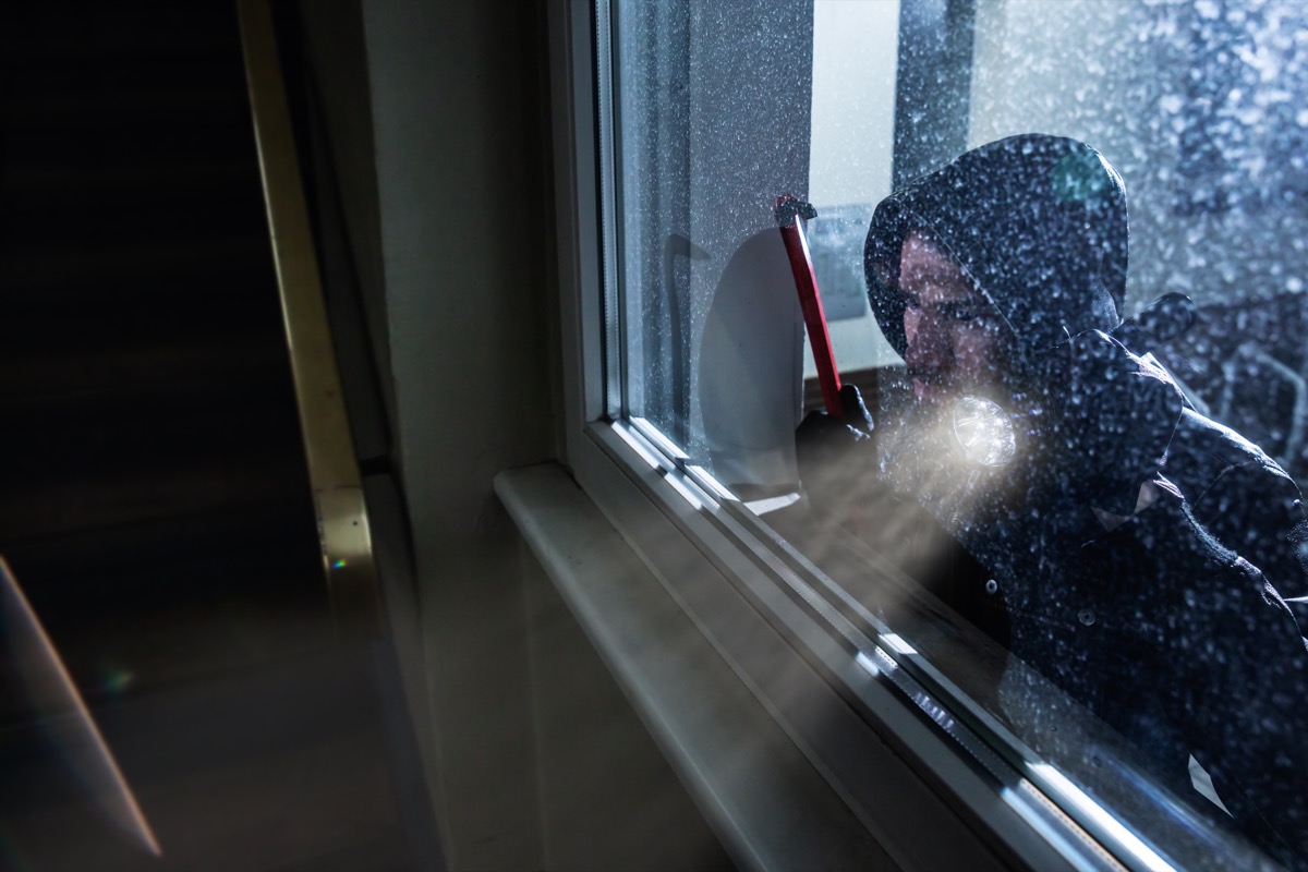 white male burglar holding crowbar and wearing hoodie peering through window of house
