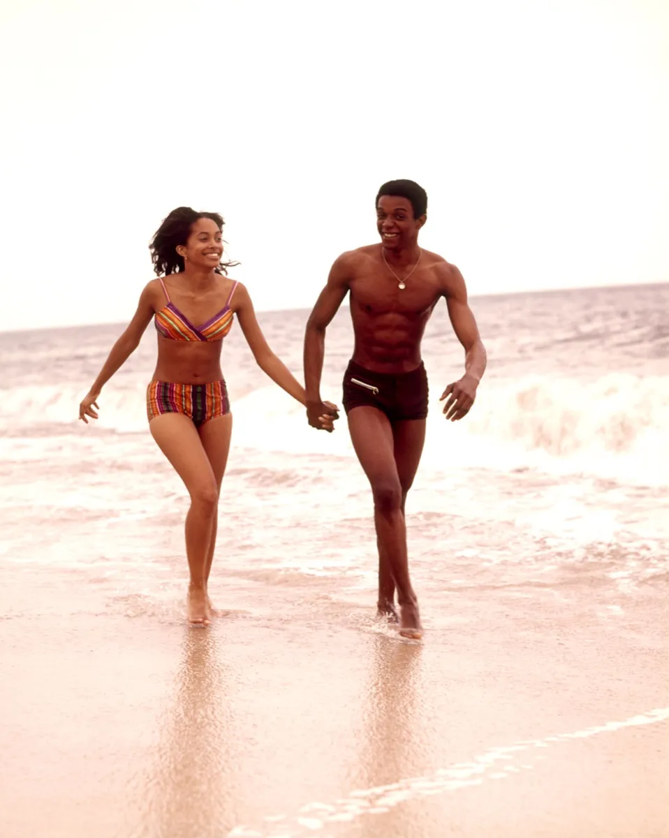 an african american couple walks hand in hand on the beach in the 1970s