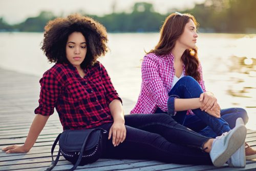 sad young black woman and young white woman sitting on the dock at a lake and not looking at each other