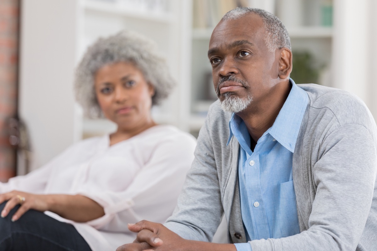 older man sitting forward on the couch while older black woman looks at home
