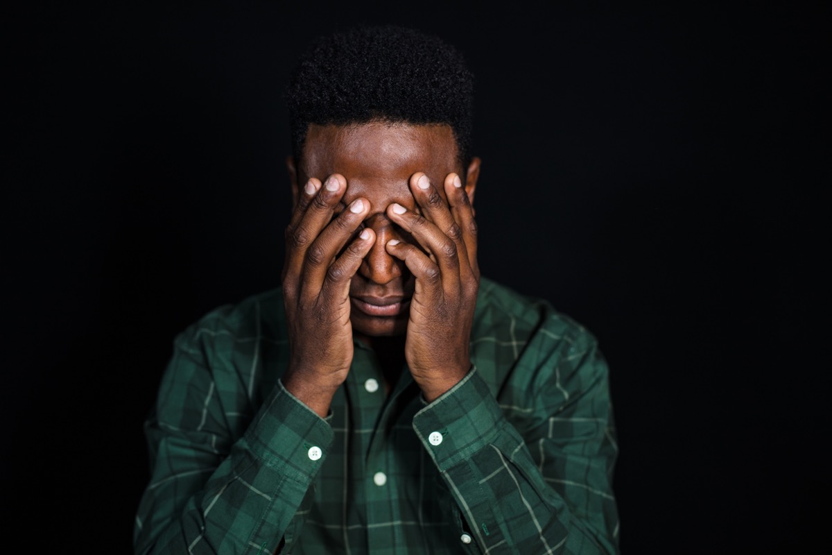 Portrait of black man with hand covering face and thinking. Male in checkered shirt looking worried on black background.