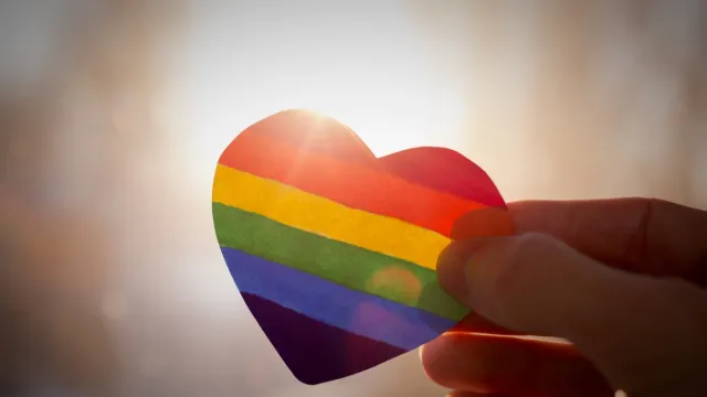 hand holds a heart painted like a LGBT flag, silhouetted against sun