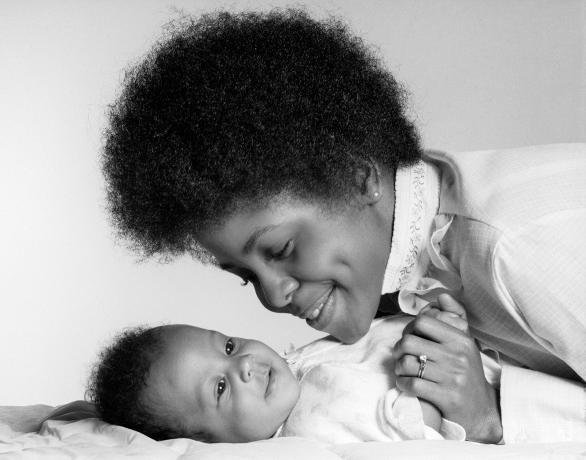 1970s black mom with afro leans over infant, holding her hand, shows how different parenting was in the 1950s