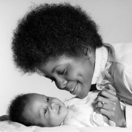 1970s black mom with afro leans over infant, holding her hand, shows how different parenting was in the 1950s