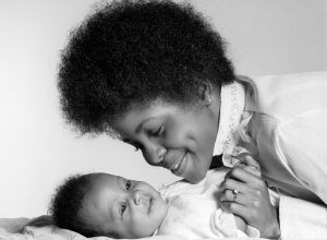 1970s black mom with afro leans over infant, holding her hand, shows how different parenting was in the 1950s