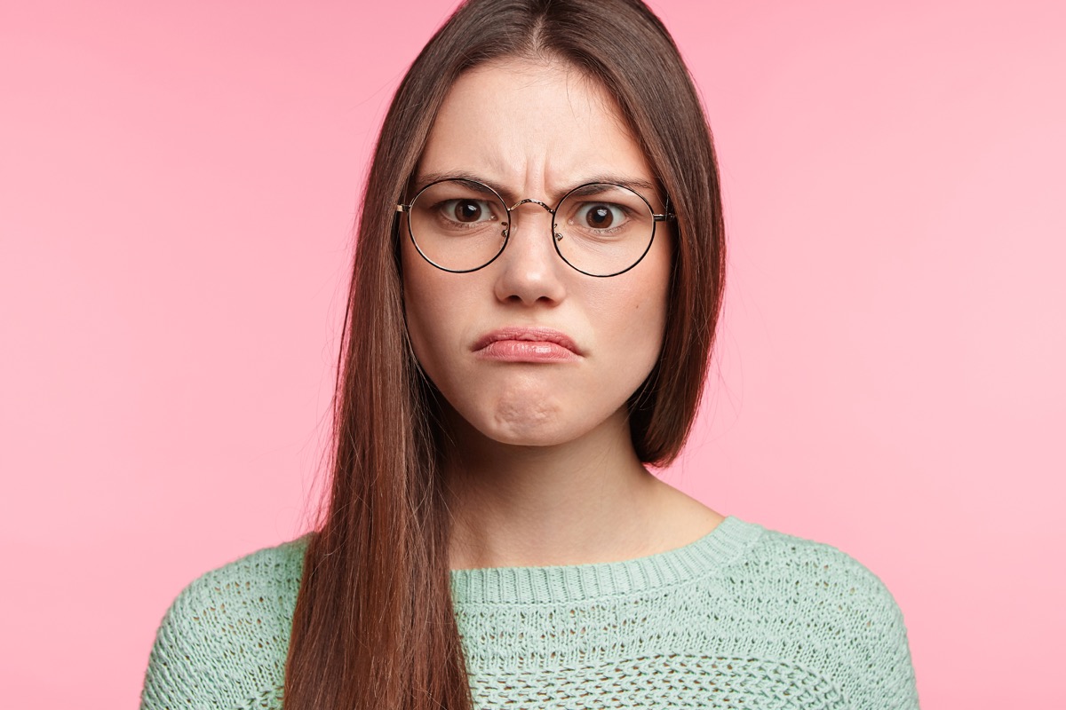 a woman frowning and wearing glasses against a light millennial pink background [backhanded compliments]