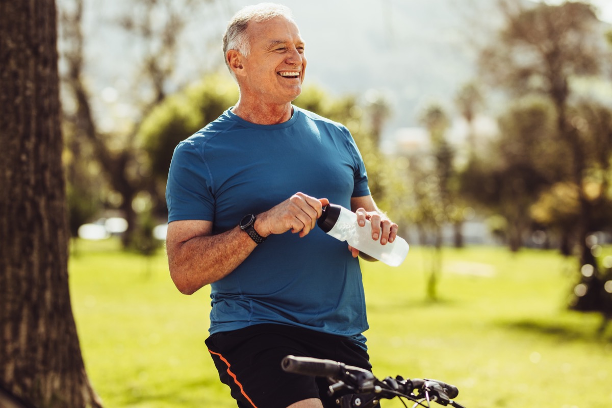 Senior man in fitness wear drinking water sitting on his bicycle. Cheerful senior fitness person taking a break during cycling in a park.