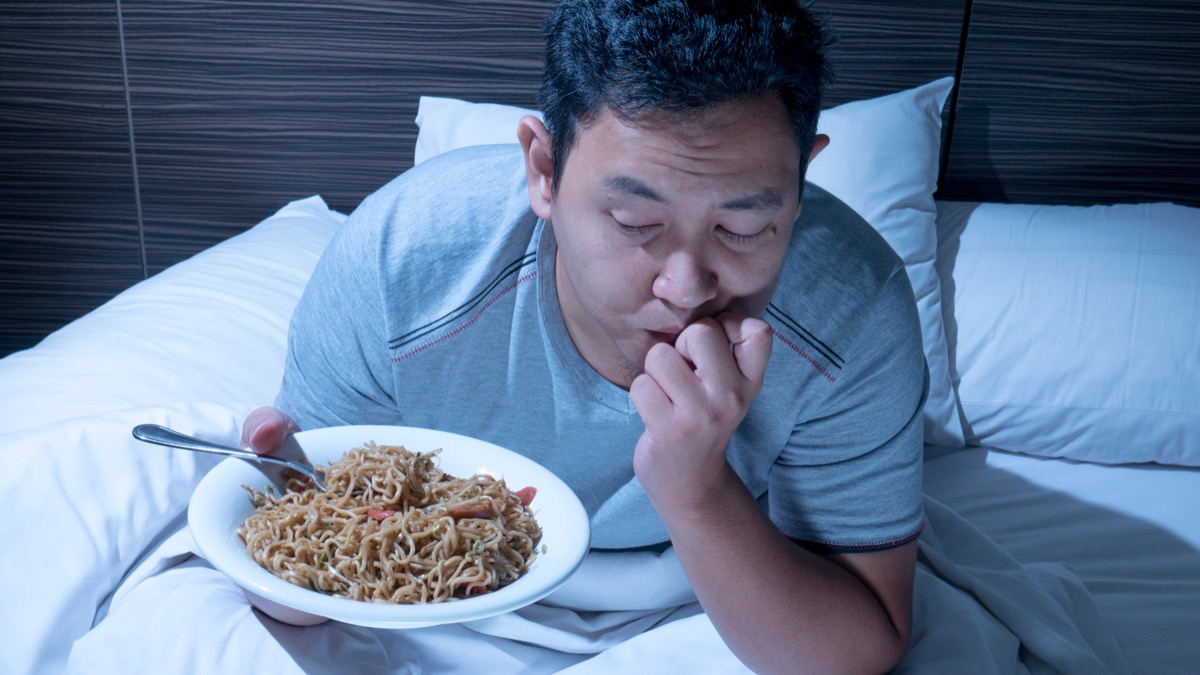 Asian man sits up in bed late at night with plate of noodles, health questions after 40