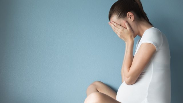 Pregnant woman holds face in hands while sitting against blue backdrop, husband left while pregnant