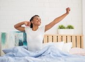 Young black woman sits up in bed in morning in white t-shirt and stretches, ways to feel amazing