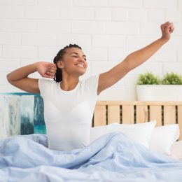 Young black woman sits up in bed in morning in white t-shirt and stretches, ways to feel amazing