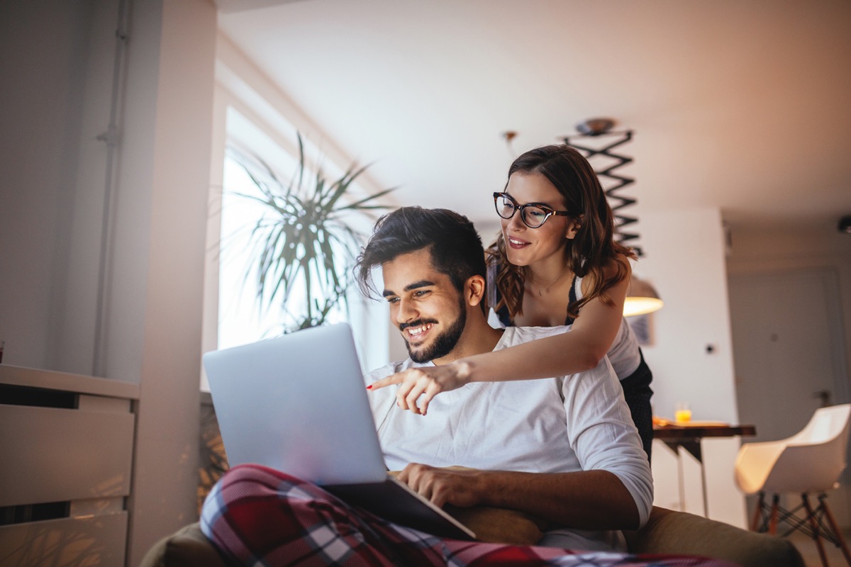 man smiling at laptop with woman pointing at screen behind him