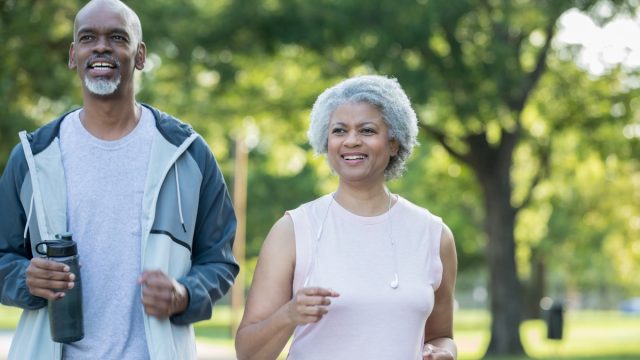 Senior adult African American couple are smiling and jogging together in public park on sunny day. Husband and wife are wearing athletic clothing.