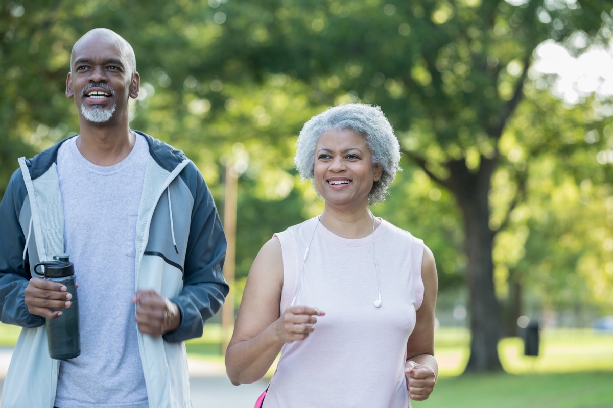 Senior adult African American couple are smiling and jogging together in public park on sunny day. Husband and wife are wearing athletic clothing.
