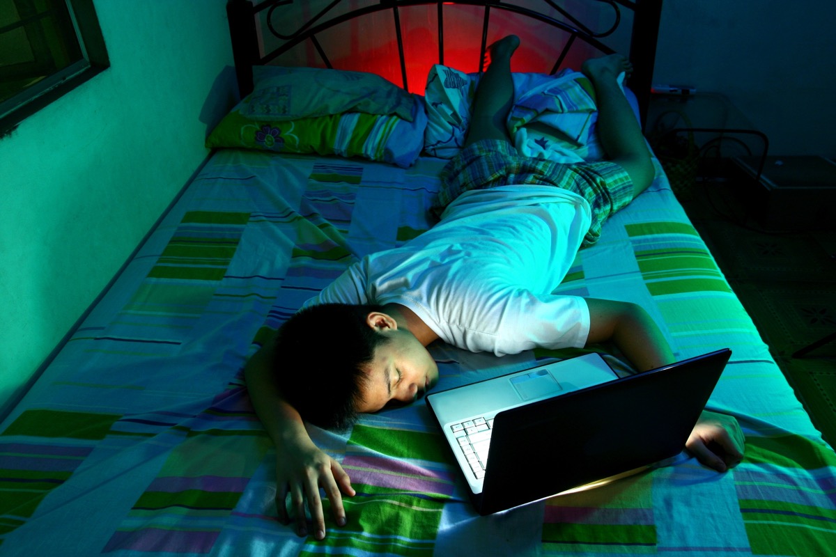 a teenager laying asleep faceup in front of a still powered on laptop doing homework