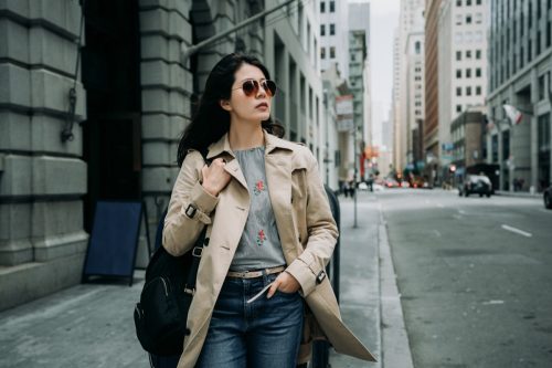 a woman wearing a tan trench coat and ray-ban sunglasses walking on a city street during a cloudy day