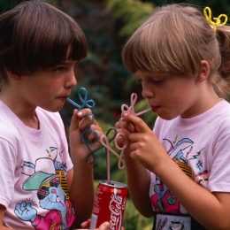 In 1980s, Two young twin sisters sip Coke from their own curly straws in an English garden, UK