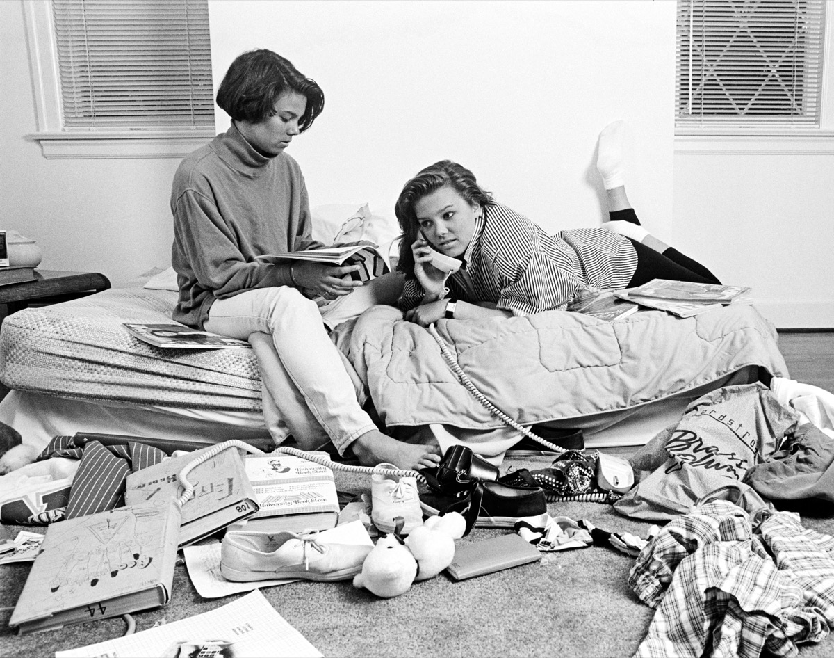 TWO TEENAGE AFRICAN AMERICAN GIRLS IN CLUTTERED BEDROOM ONE TALKING ON THE PHONE ONE READING MAGAZINE