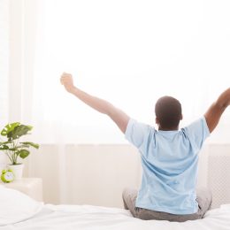 Black man pictured from behind stretching in bed in the morning, wearing a blue shirt, savory breakfast leads to success study