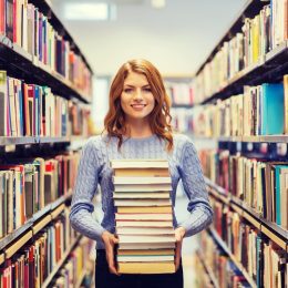 Young woman standing in library aisle holding stack of books