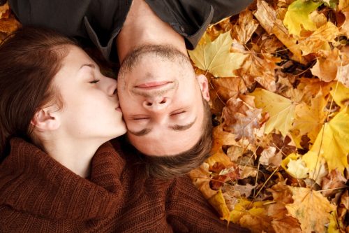 Frau küsst Mann auf die Wange, während sie im Laub liegt - Romantische Ideen für ein Date im Herbst