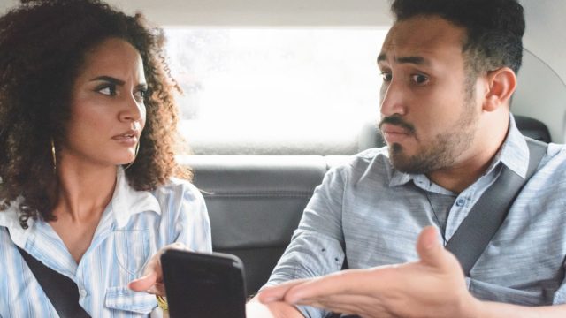 young couple arguing in the backseat of a car while the man points to his phone defensively