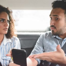 young couple arguing in the backseat of a car while the man points to his phone defensively