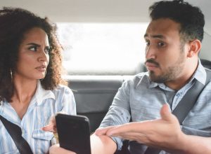 young couple arguing in the backseat of a car while the man points to his phone defensively