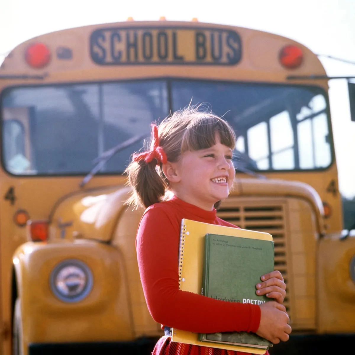 1970s SMILING ELEMENTARY SCHOOL GIRL STANDING FRONT OF BUS CARRYING BOOKS SMILING