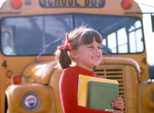 1970s SMILING ELEMENTARY SCHOOL GIRL STANDING FRONT OF BUS CARRYING BOOKS SMILING
