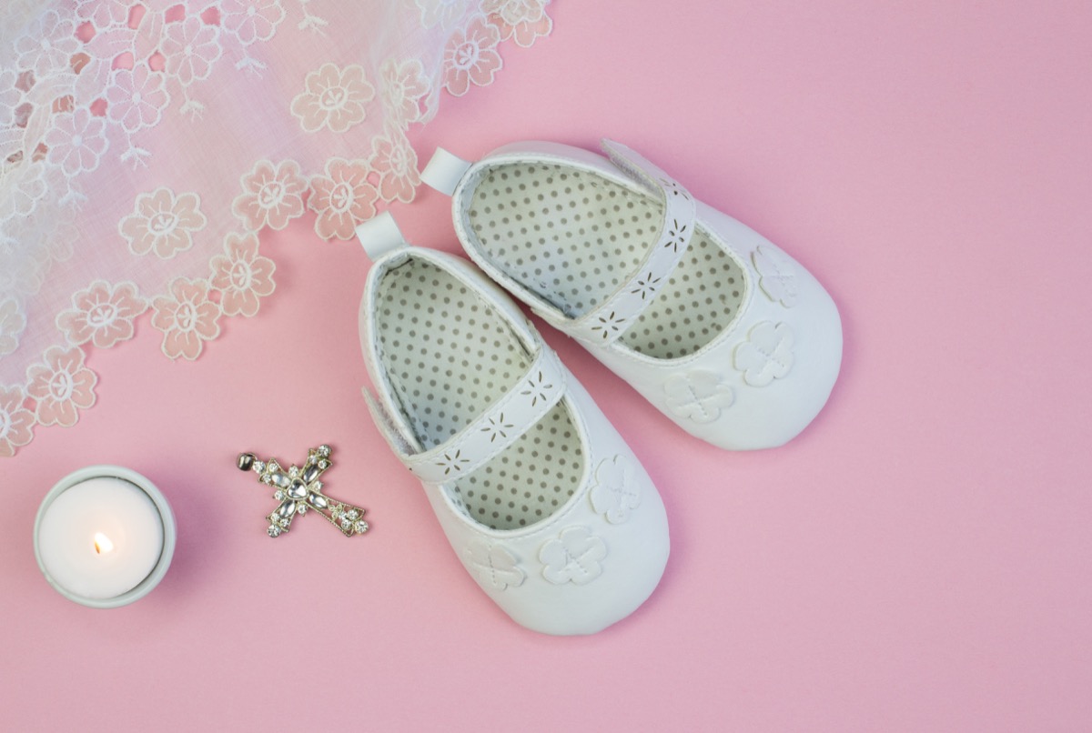 Pair of white baby booties on pink background with lace christening dress and candle