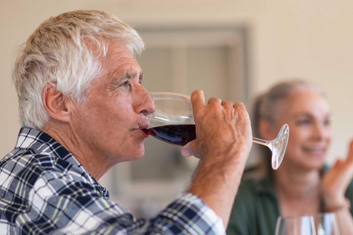Happy senior man drinking a glass of red wine during lunch. Old man enjoying wine with friends in background. Closeup face of active and healthy senior man tasting wine.