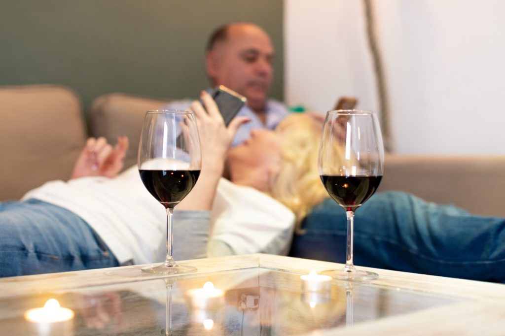 Two glasses of red wine on table with senior couple relaxing in background on sofa with smartphones in their hands. (Two glasses of red wine on table with senior couple relaxing in background on sofa with smartphones in their hands., ASCII, 116 compon