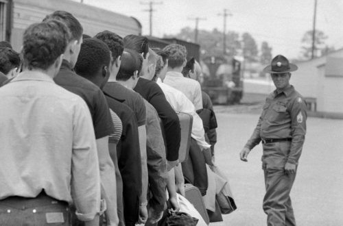 Black and white photo of Army recruits in street clothes waiting to board bus with officer in the background