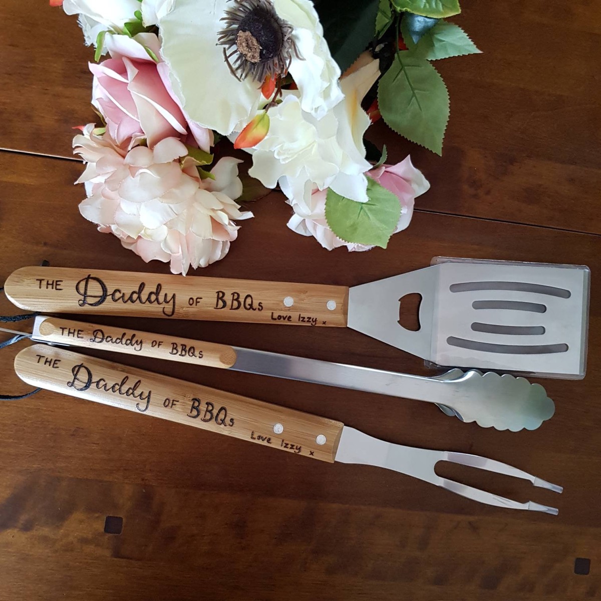 set of three barbecue tools with engraved wooden handles on wood table next to flowers
