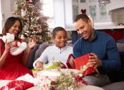 young black man sitting on couch with black woman and young boy opening a christmas present in a red box
