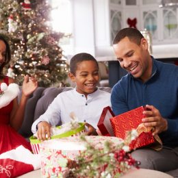 young black man sitting on couch with black woman and young boy opening a christmas present in a red box