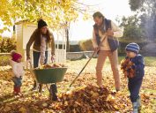 black mother, father, and two little girls raking leaves into green wheelbarrow on a fall day