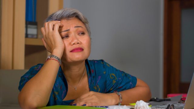 frustrated asian woman with short gray hair leans head on hand while sitting at desk