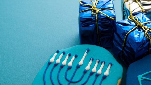 blue hanukkah cookie with menorah on it next to two small gifts wrapped in blue metallic paper and gold ribbon, hanukkah gifts
