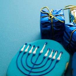 blue hanukkah cookie with menorah on it next to two small gifts wrapped in blue metallic paper and gold ribbon, hanukkah gifts
