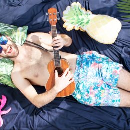 man in sugnlasses and boxers playing ukulele in bed on blue sheets