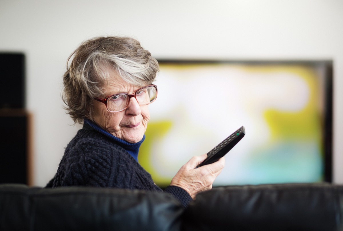 An irritated senior woman turns from the television she has been watching, holding the remote control.