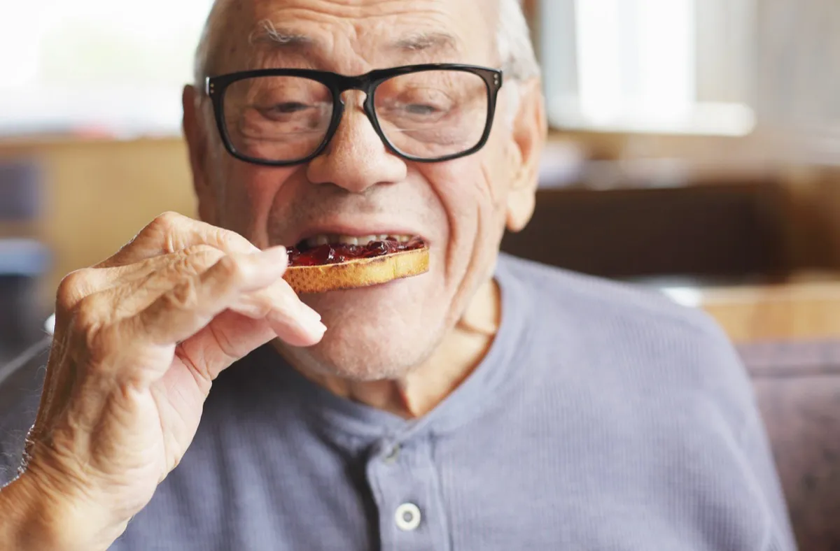 A senior man enjoys a bite of his toasted bread spread with sweet jelly jam preserves during breakfast at a restaurant.