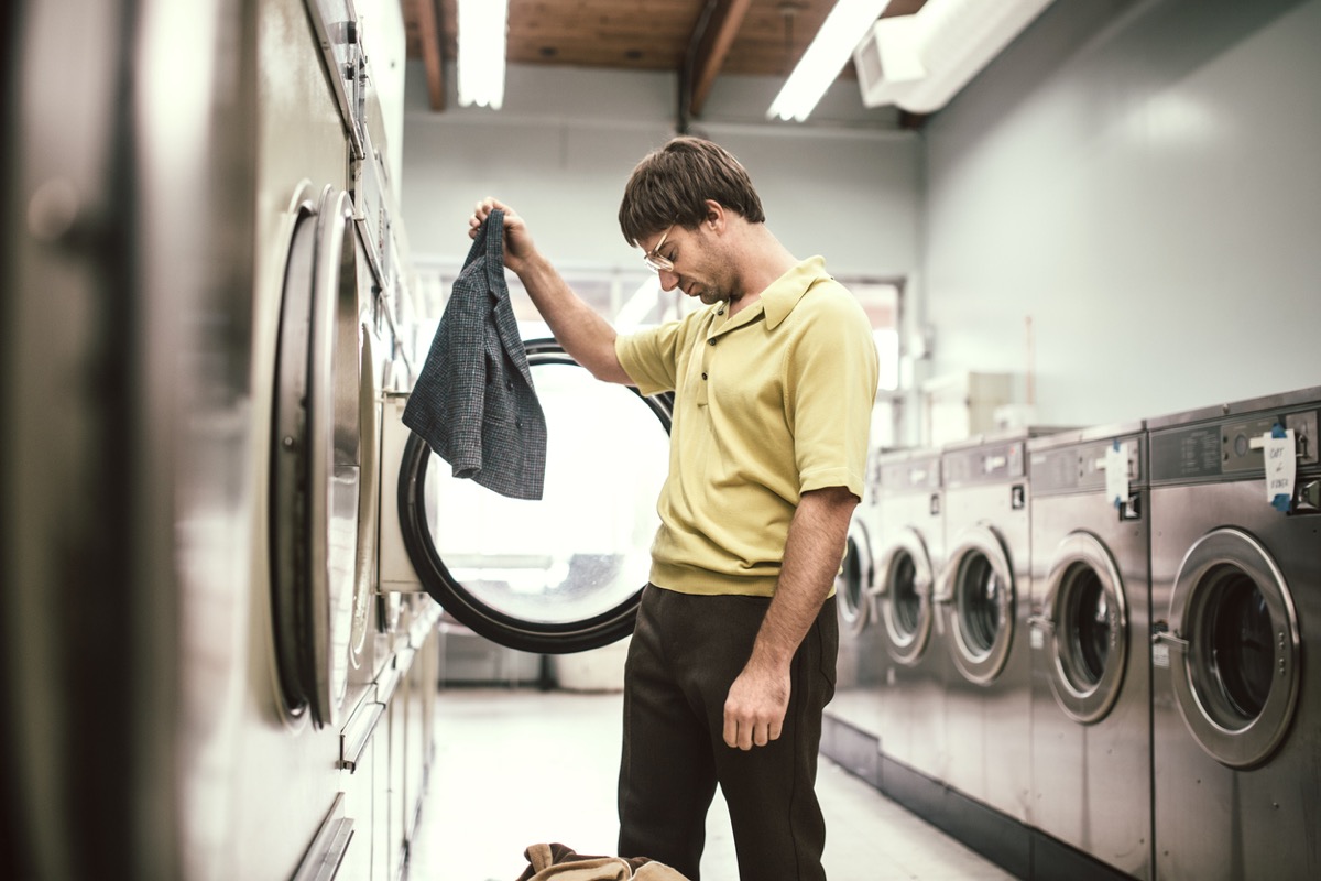 A man in 1980's style does his laundry at an old school laundromat. He holds up his jacket which has shrunken to a ridiculously small size while drying in the dryer, his head hanging down in sadness and disappointment.